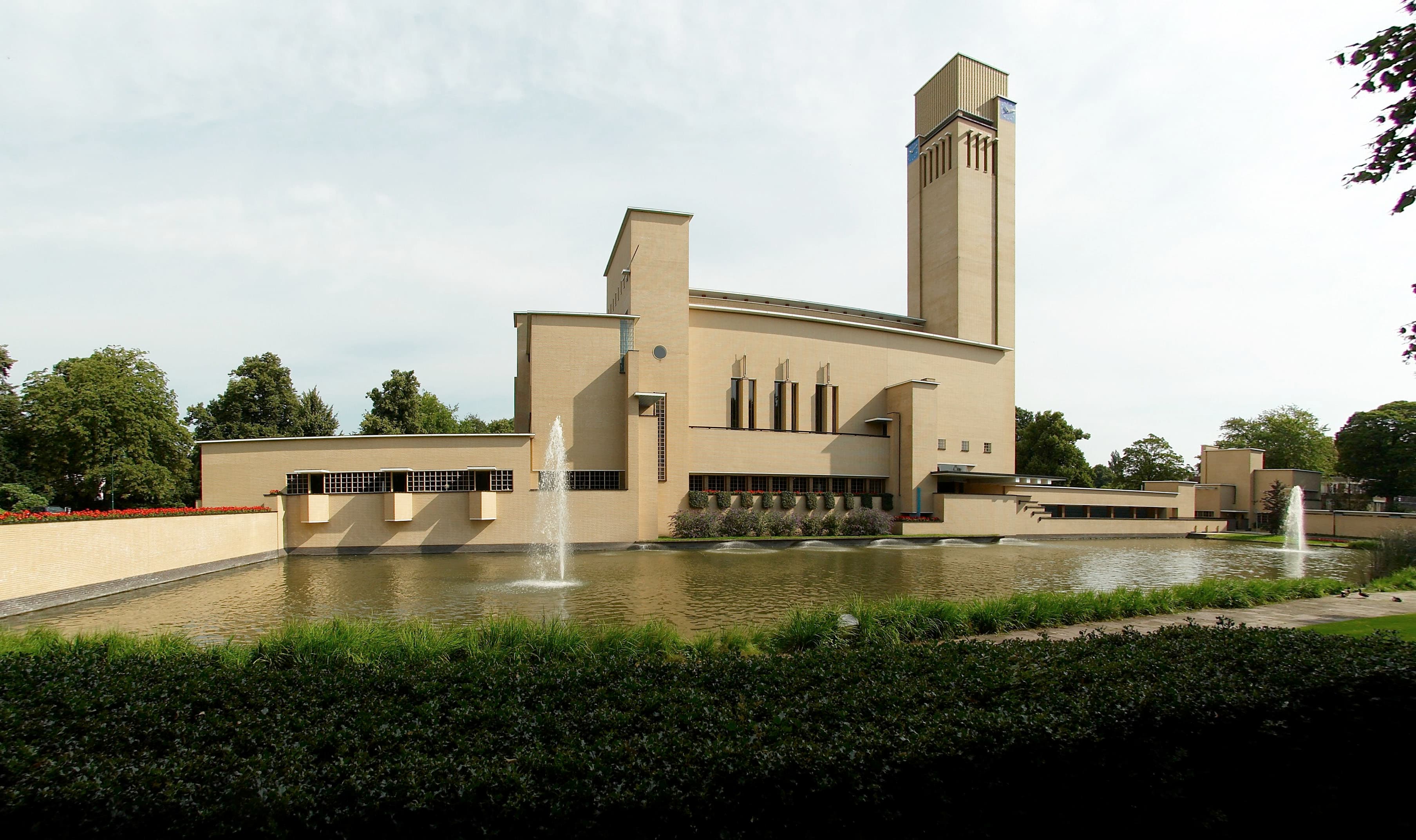 Het raadhuis van Hilversum in gele baksteen, met een hoge toren en een vijver met fonteinen op de voorgrond.