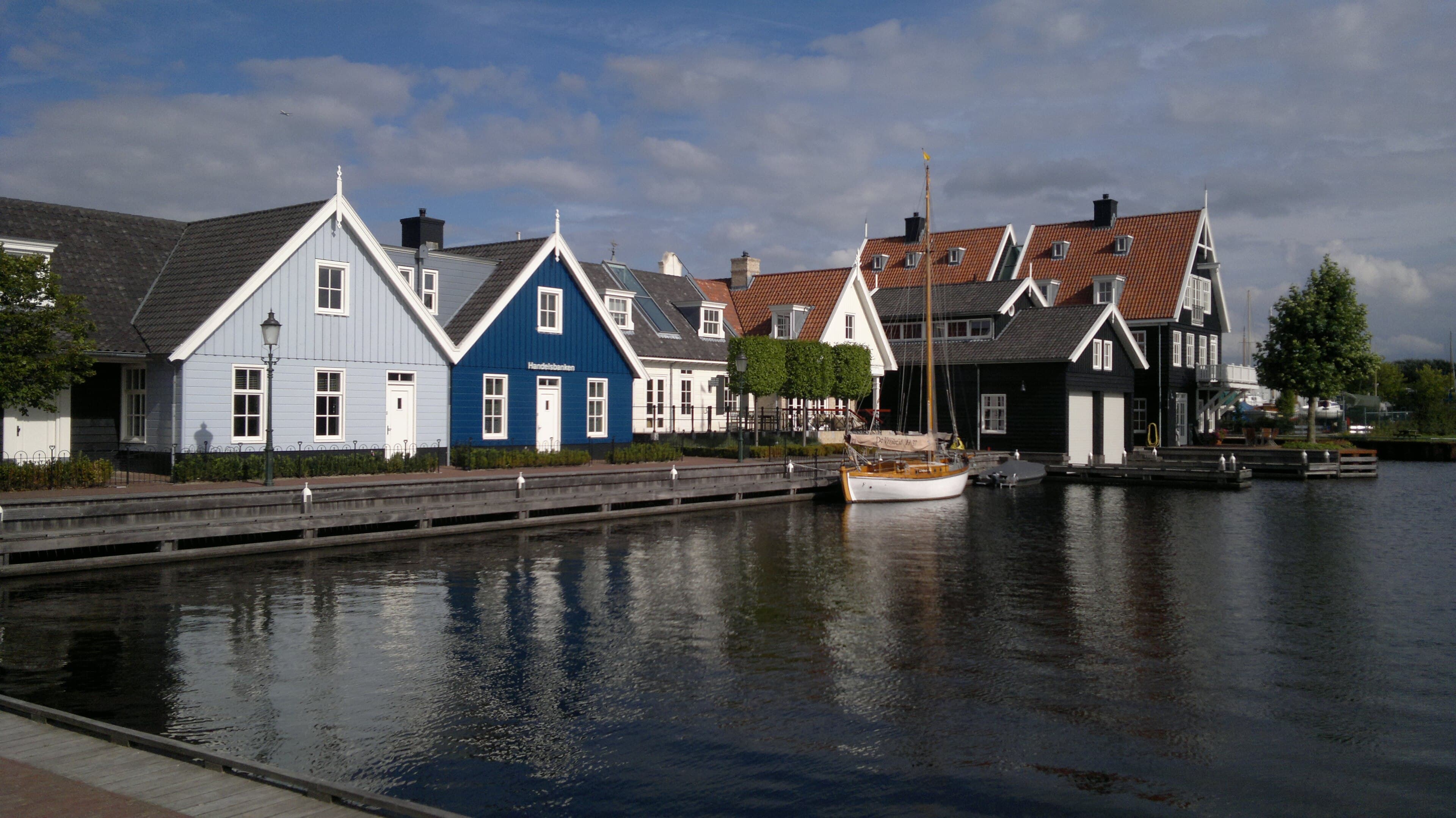 Houten huizen in blauw, wit en zwart aan de kade van het Nautisch Kwartier in Huizen, met een houten zeilboot die weerspiegelt in het havenwater