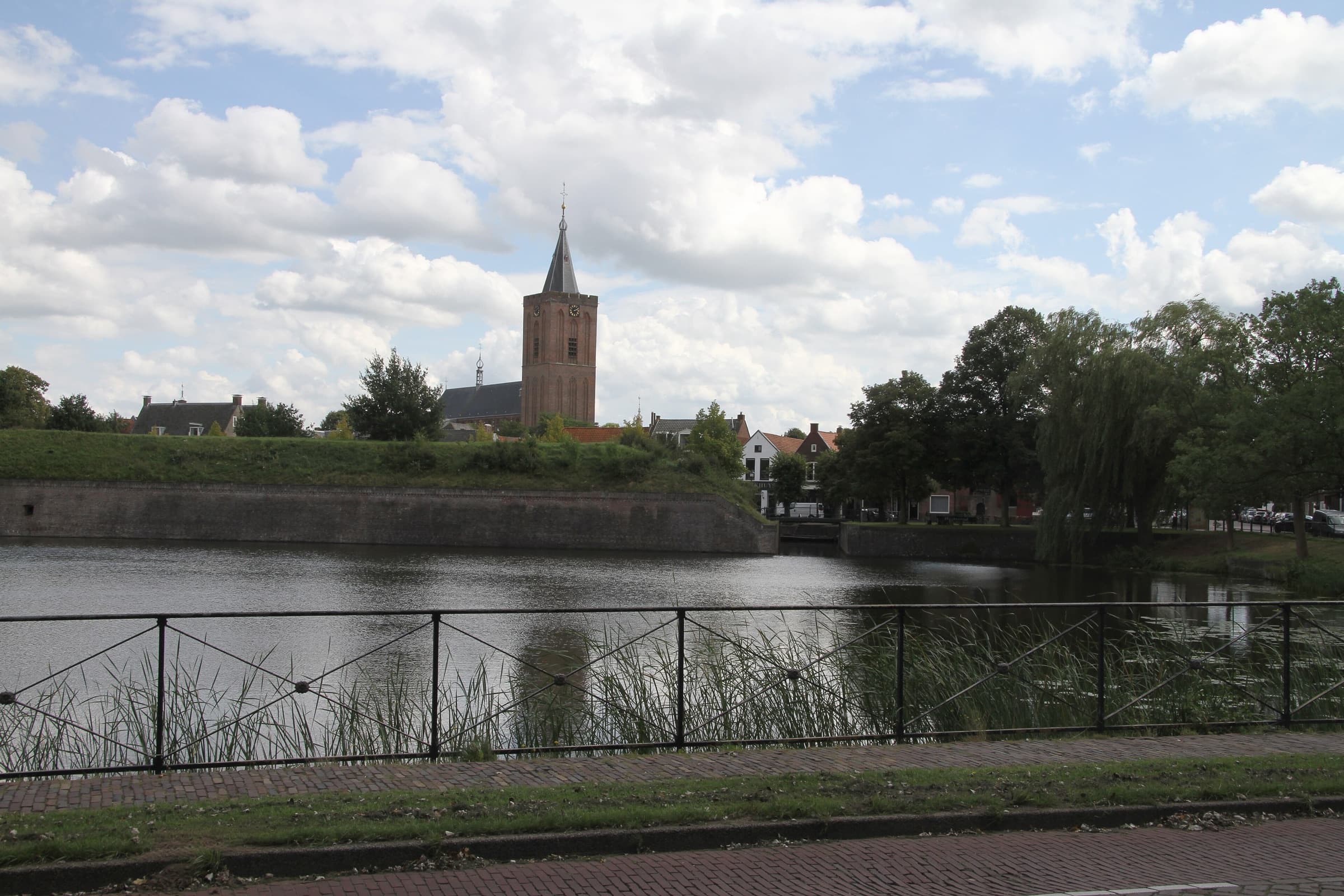 De vestinggracht van Naarden met een bakstenen wal, daarachter de toren van de Grote Kerk en lage huizen.