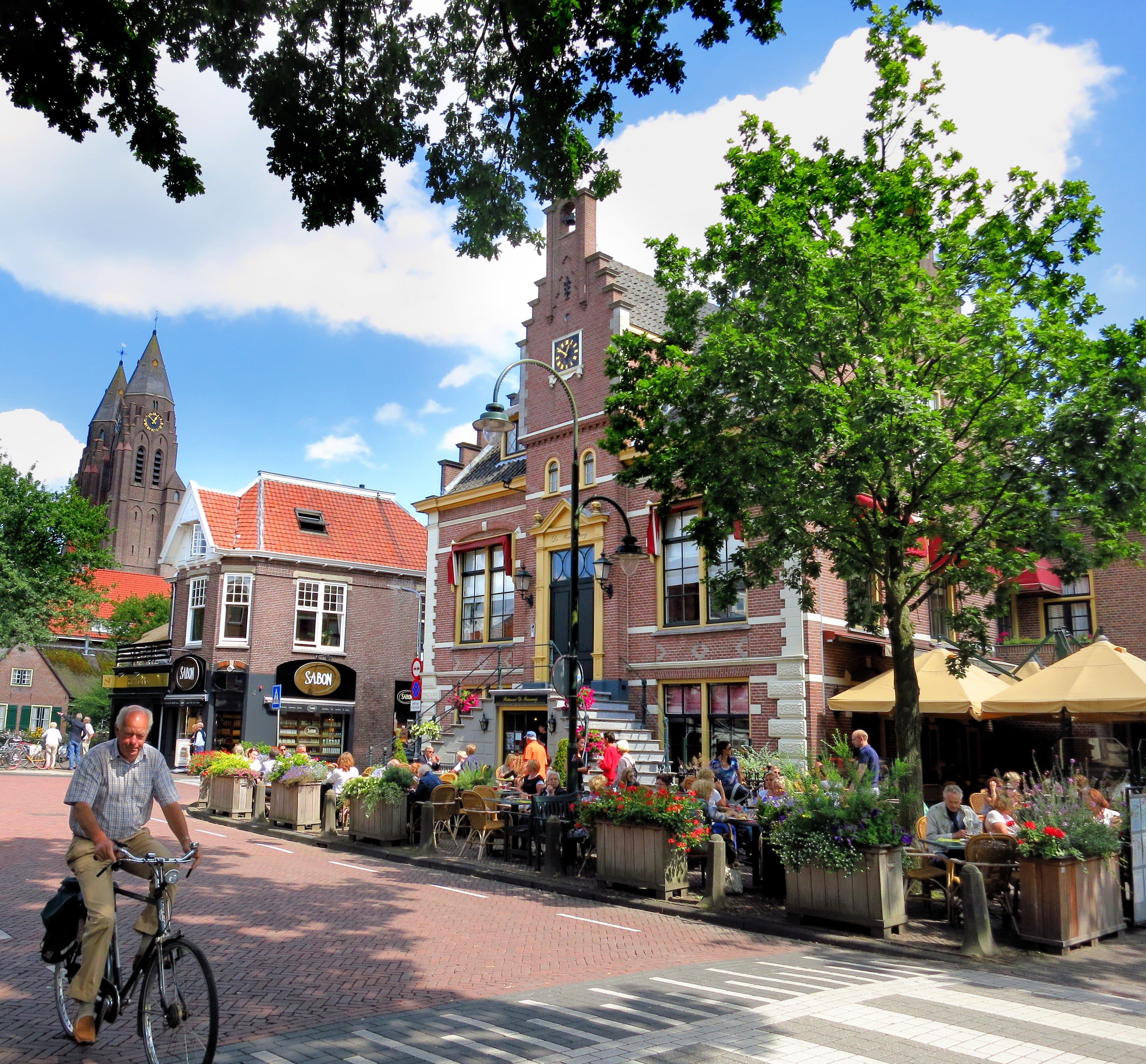 Zonnig plein in Laren met een fietser op de voorgrond, een terras met parasols en mensen aan tafeltjes, en op de achtergrond de torens van de Sint-Jansbasiliek